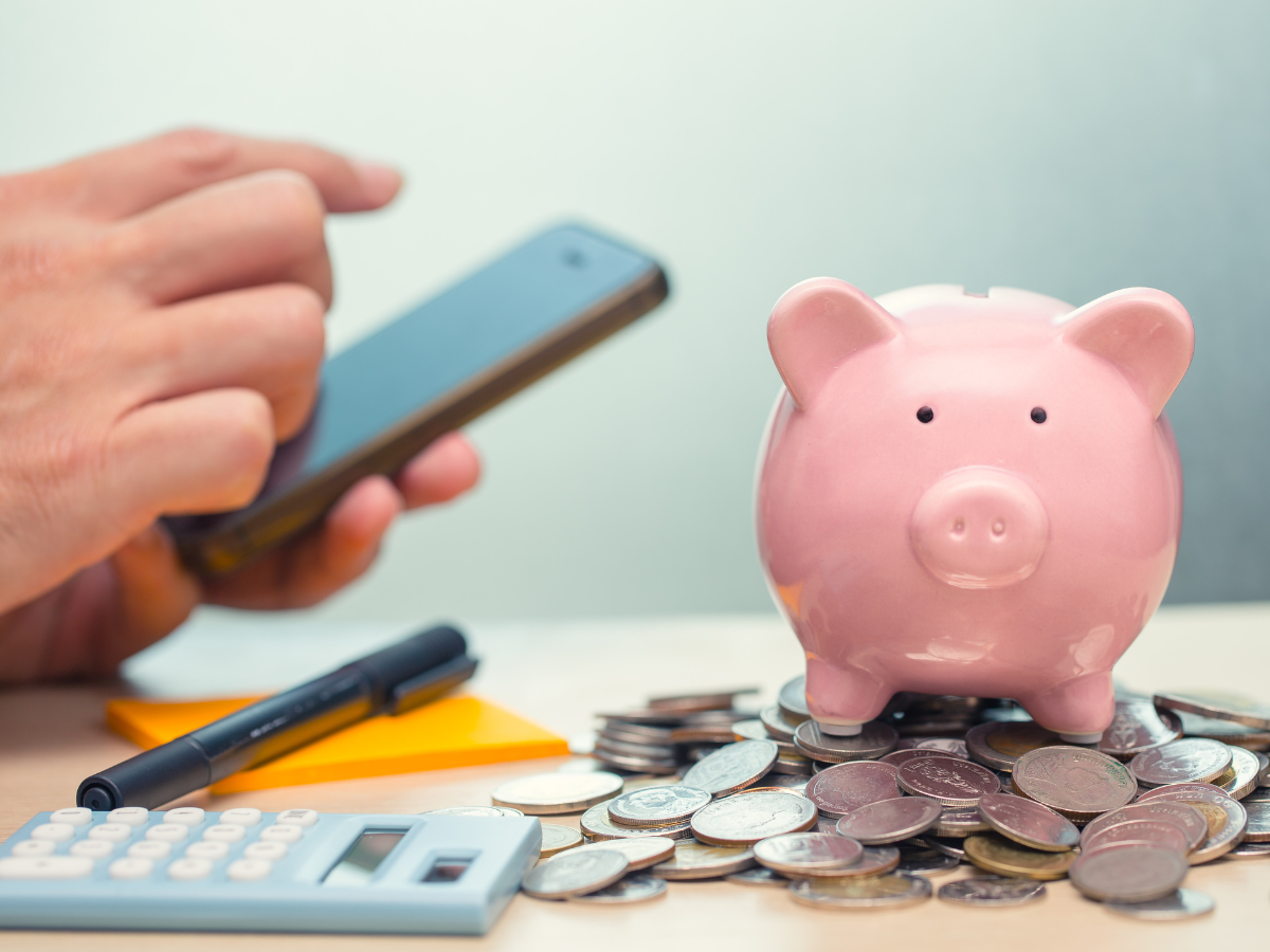 A piggy bank sitting atop of coins with a persons hand holding a phone to signify financial apps