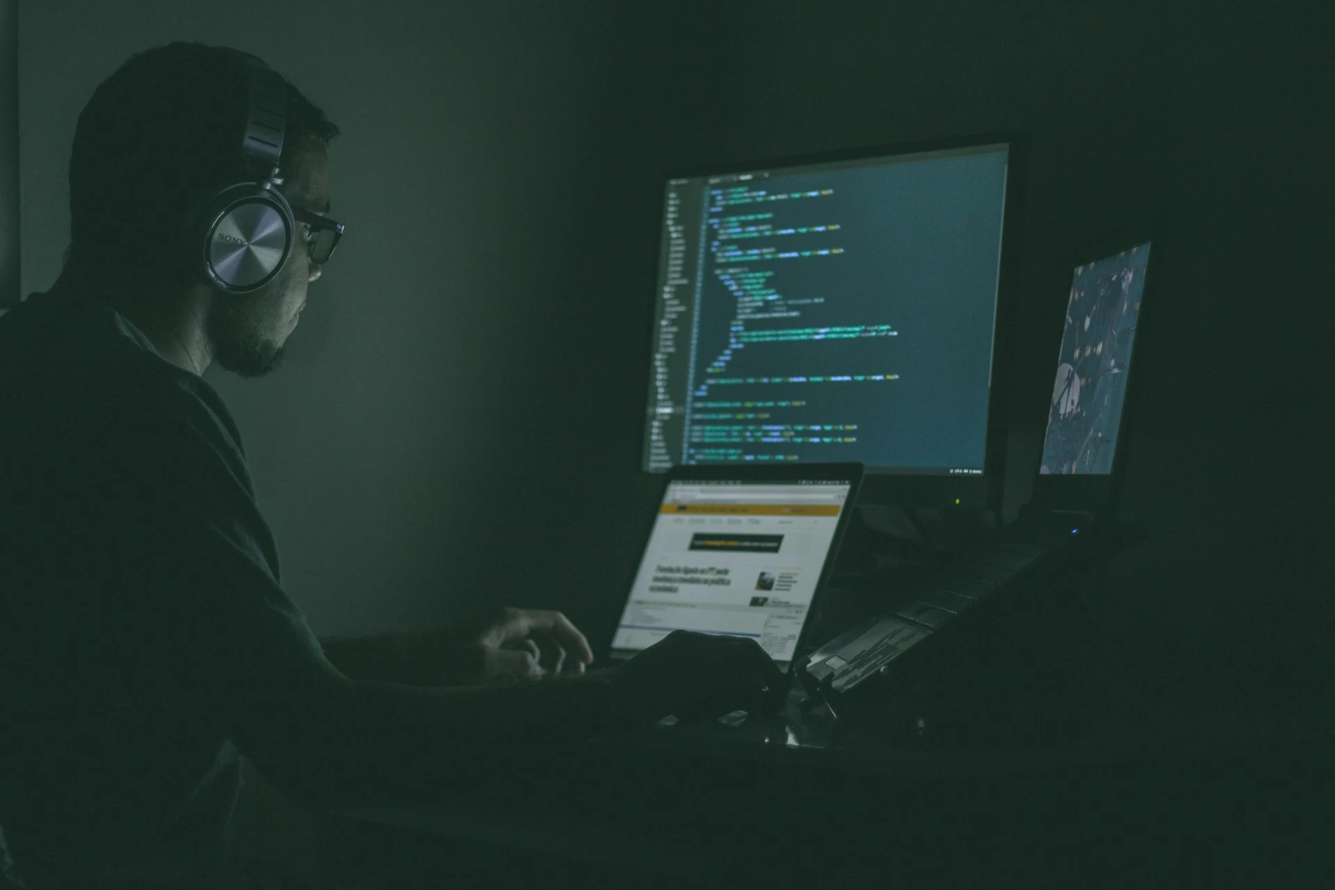 A man wearing headphones sits at a desk with two monitors, engrossed in his work.