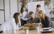 A diverse group of professionals collaborates around a laptop at a modern office table, with sticky notes and documents scattered around.