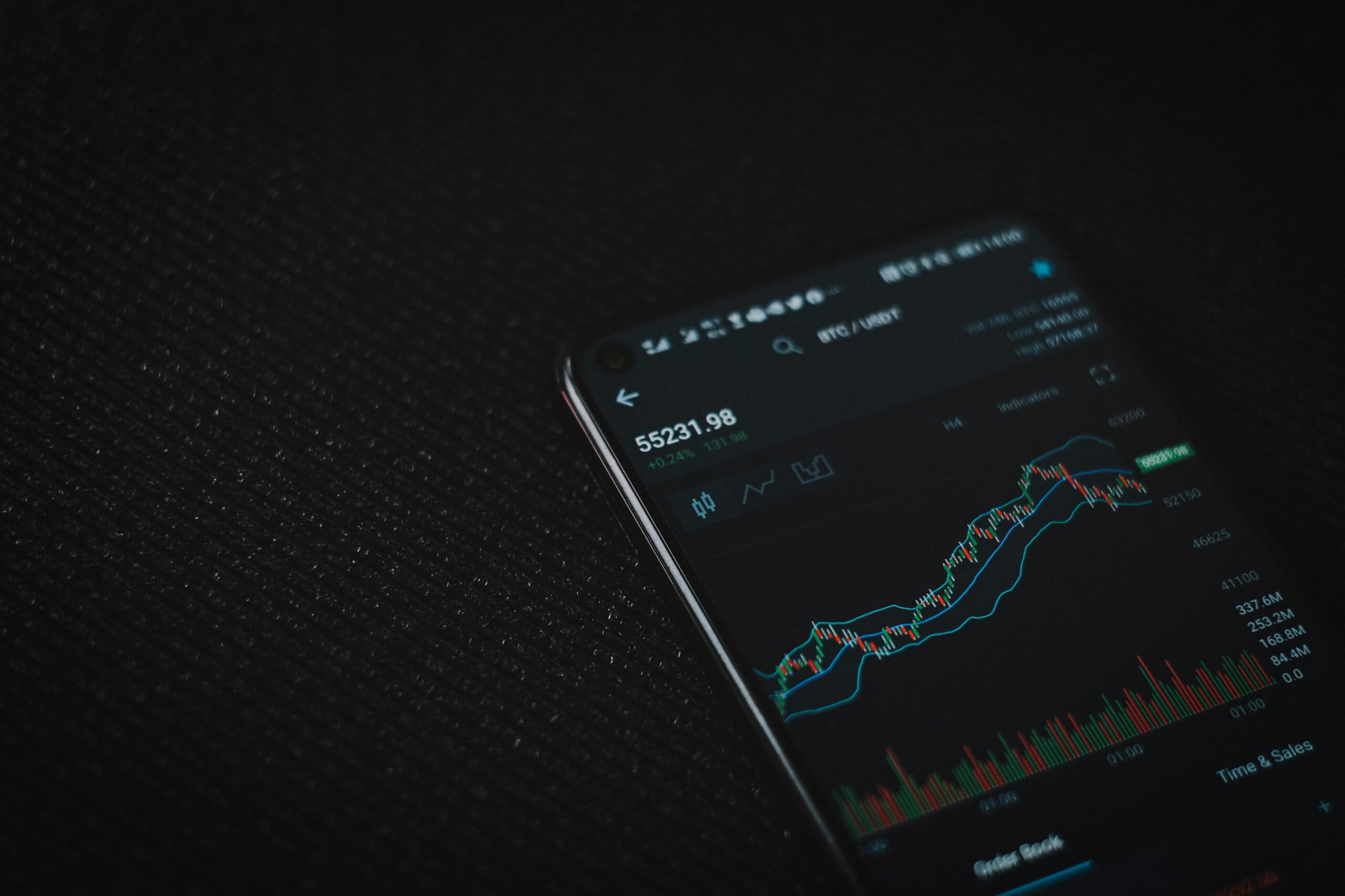 A smartphone displaying a cryptocurrency price chart and trading interface on a dark textured surface. The screen shows a rising candlestick chart with market indicators and trading volume, creating a dark, technology-focused financial atmosphere.